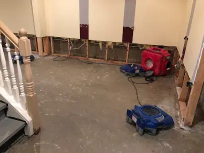 Basement with lower drywall cut away, a dehumidifier and air movers drying the floor near the stairs in a West Chester home